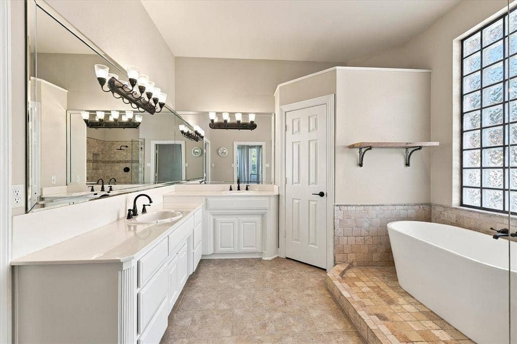 Dual vanity with white cabinetry and integrated sinks, a freestanding tub, tiled bathing area, large glass block window, and extensive wall-mounted mirrors