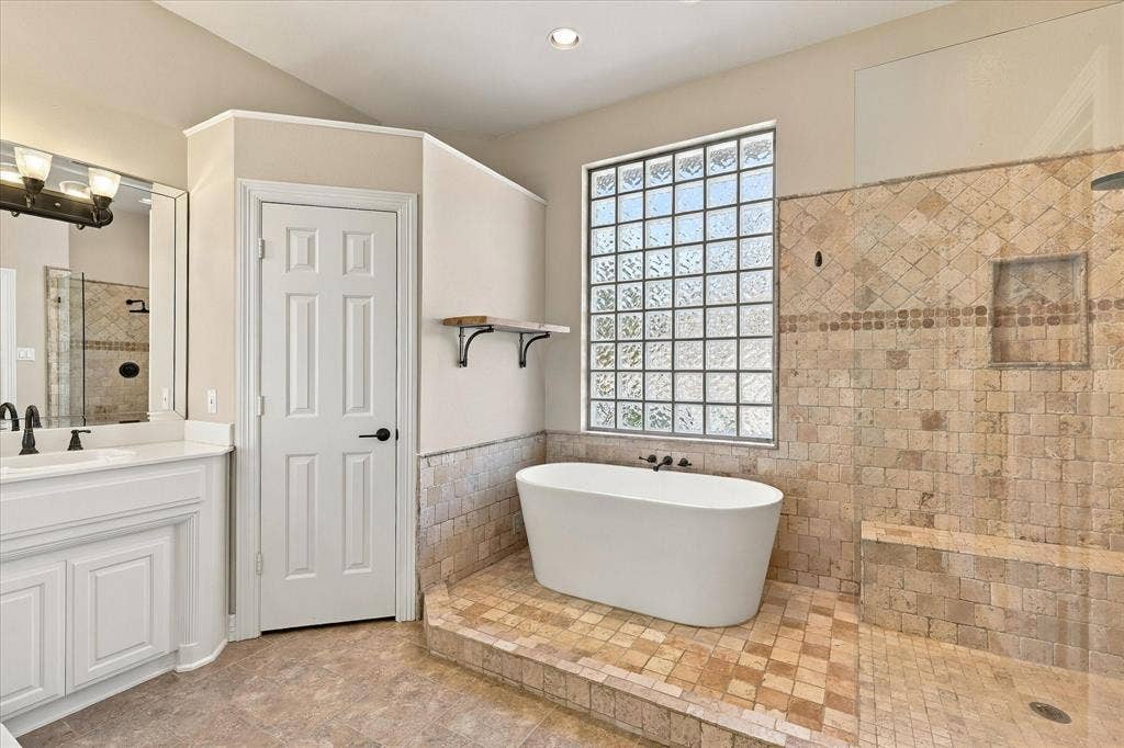 Freestanding bathtub, glass block window, tiled shower with built-in bench, vanity with undermount sink, and dark bronze fixtures