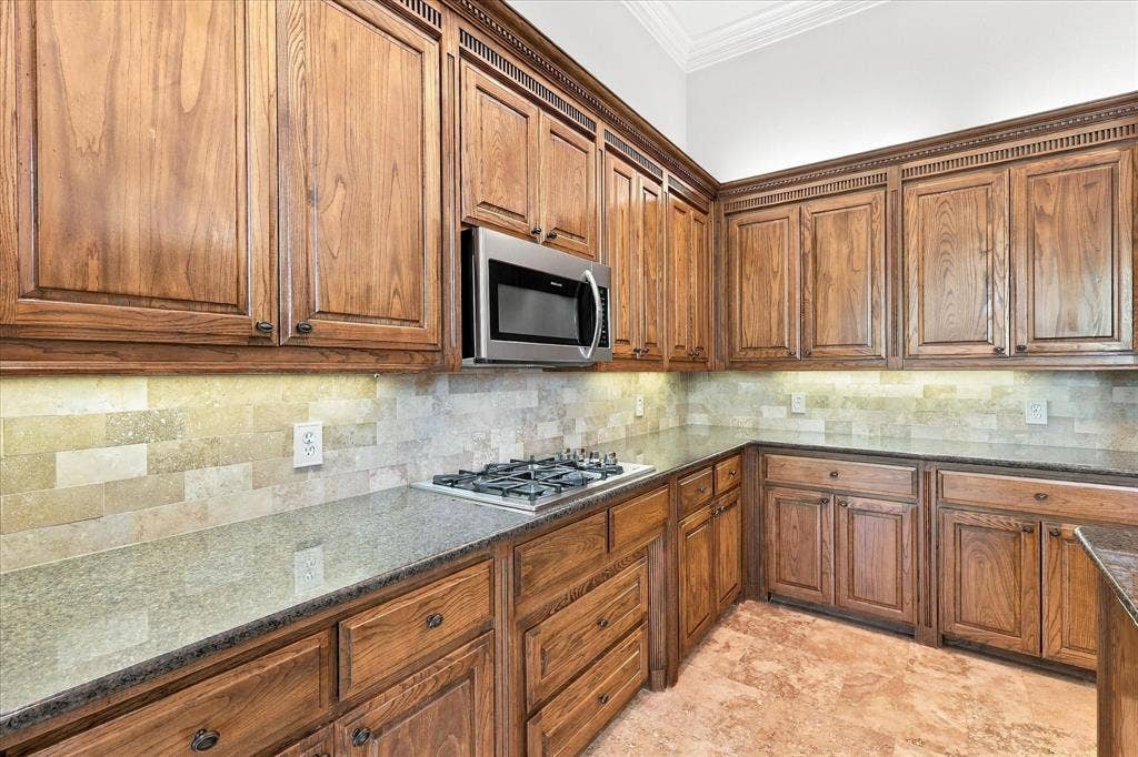 Extensive wood cabinetry, stainless steel microwave, gas cooktop, stone tile backsplash, and light-toned flooring