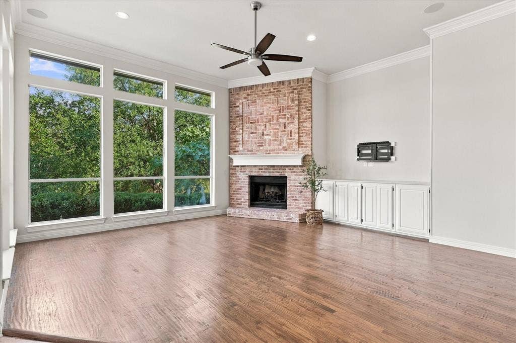 Hardwood-finish flooring, a brick fireplace with a white mantel, white built-in cabinetry, an angular window array, and a ceiling fan with integrated lighting