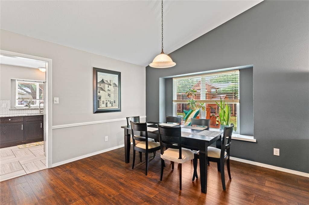 Vaulted ceiling with pendant lighting, wood-finish flooring, two-tone wall treatment, and a large window with blinds