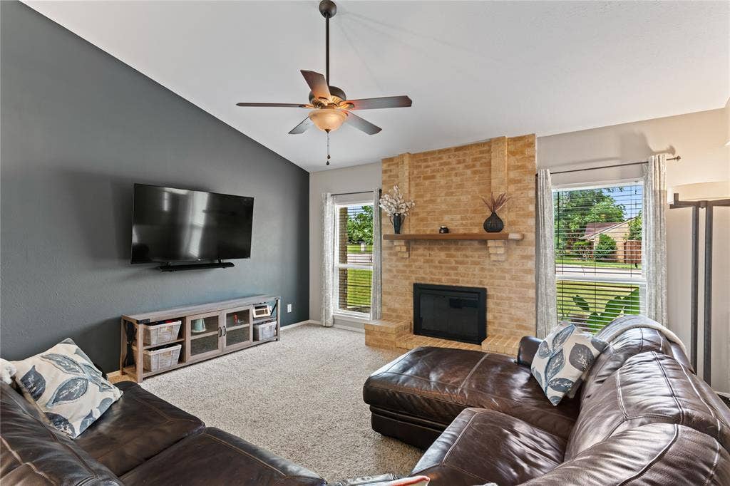 Vaulted ceiling, ceiling fan with light fixture, brick fireplace with wood mantel, large windows with blinds, and wood look laminate flooring