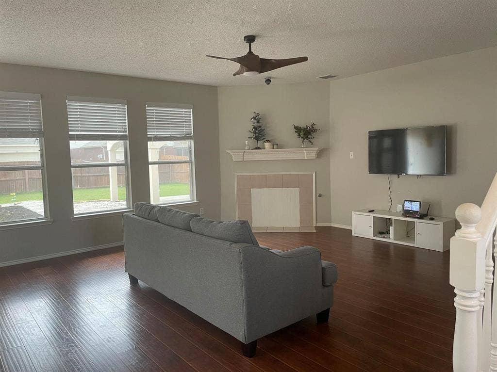 Living area featuring wood-finish flooring, a fireplace with a mantel, triple windows, and a ceiling fan