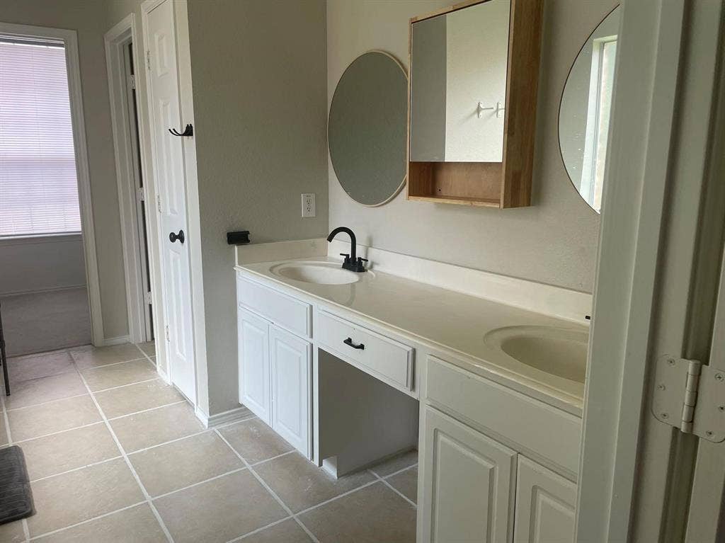 Dual-sink vanity with matte black faucets, light-toned countertop, round wall mirrors, and built-in medicine cabinet, complemented by tile flooring