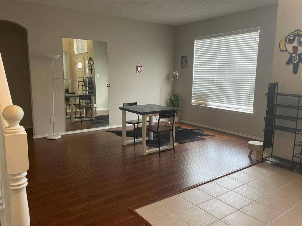 Living area featuring wood-finish flooring, floor-to-ceiling wall mirror, ceramic tile inlay at entryway, large window with horizontal blinds, and staircase with wooden balustrade