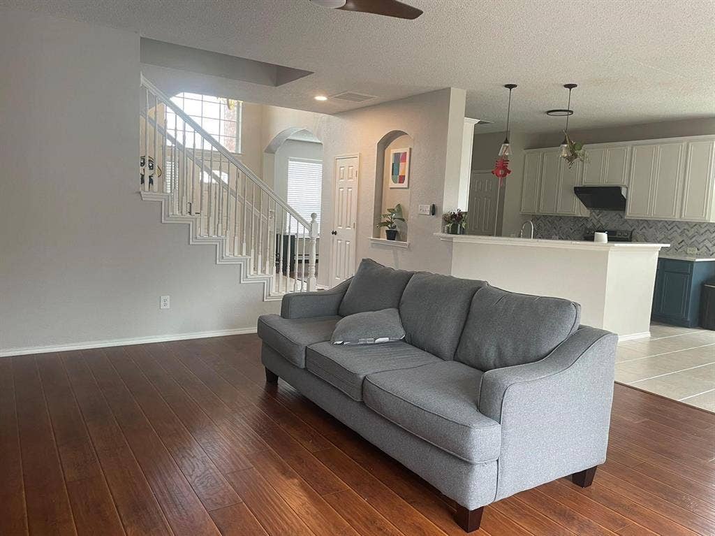 Open-concept interior featuring wood-finish flooring, a white staircase with decorative metal balusters, an arched wall niche, and a kitchen equipped with a peninsula and herringbone-patterned tile backsplash