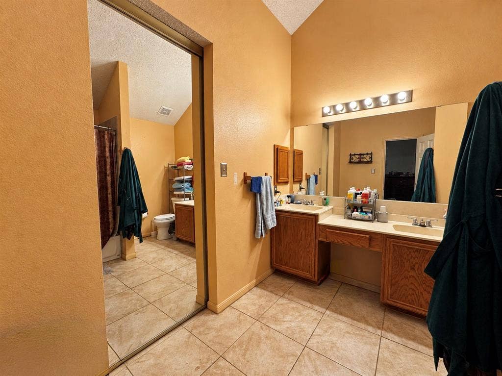 725 Andalusia Trail, DeSoto, TX 75115 Vaulted ceiling bathroom featuring a dual vanity with oak-finish cabinetry, integrated sinks, and an expansive wall-mounted mirror