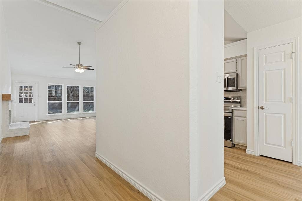 7440 Bear Lake Drive, Fort Worth, TX 76137 Vaulted ceiling with a ceiling fan, recessed windows, wood-finish flooring, white painted brick fireplace, and a white kitchen with stainless steel appliances