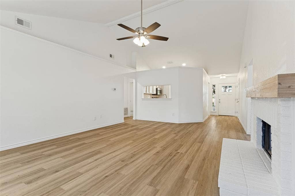 7440 Bear Lake Drive, Fort Worth, TX 76137 Vaulted ceiling with ceiling fan, wood-finish flooring, white brick fireplace with wood mantel, built-in kitchen pass-through, and white paneled entry door with sidelights
