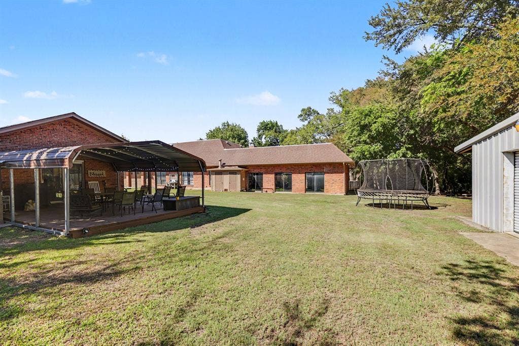 26 Brookhaven Drive, Lucas, TX 75002 Expansive backyard with a covered patio deck, brick exterior, multiple sliding glass doors, and a detached corrugated metal structure