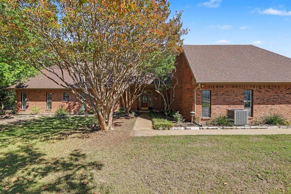 26 Brookhaven Drive, Lucas, TX 75002 Brick exterior with a brown shingle roof, mature trees, and a concrete walkway