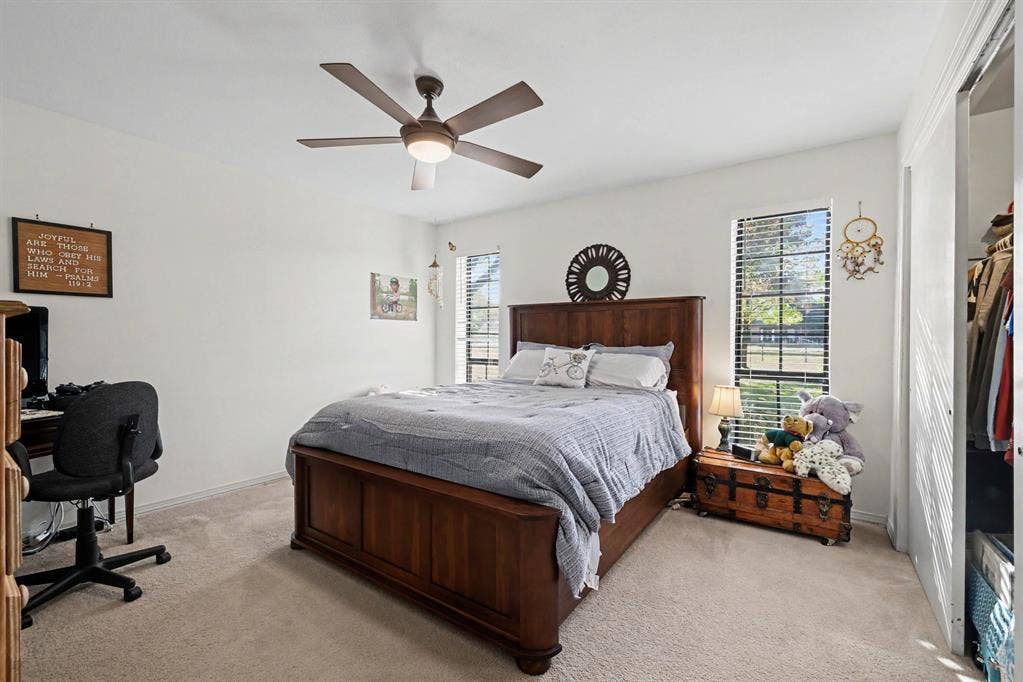 26 Brookhaven Drive, Lucas, TX 75002 Spacious primary bedroom featuring a wood-finish bed frame, ceiling fan with integrated lighting, neutral carpet flooring, and natural light from multiple windows
