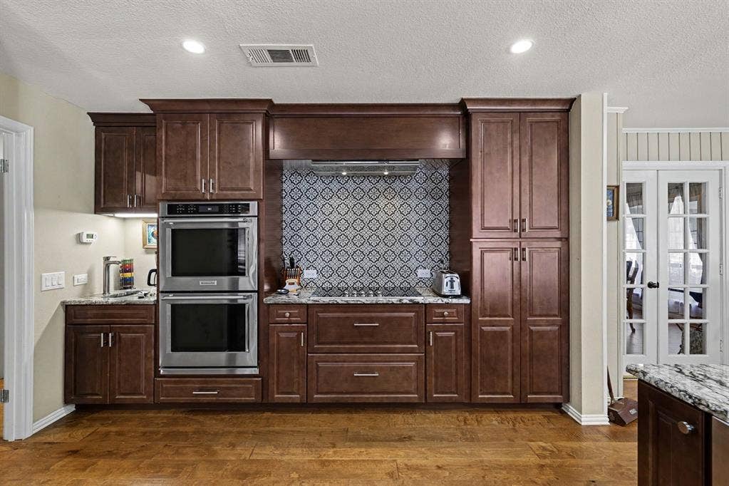 26 Brookhaven Drive, Lucas, TX 75002 Wood-finish flooring, dark wood cabinetry, stainless steel double wall ovens, patterned tile backsplash, and light-toned countertops