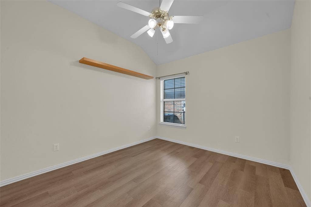 Vaulted ceiling, ceiling fan with light fixture, wood-finish flooring, built-in wall shelving, and window with blinds
