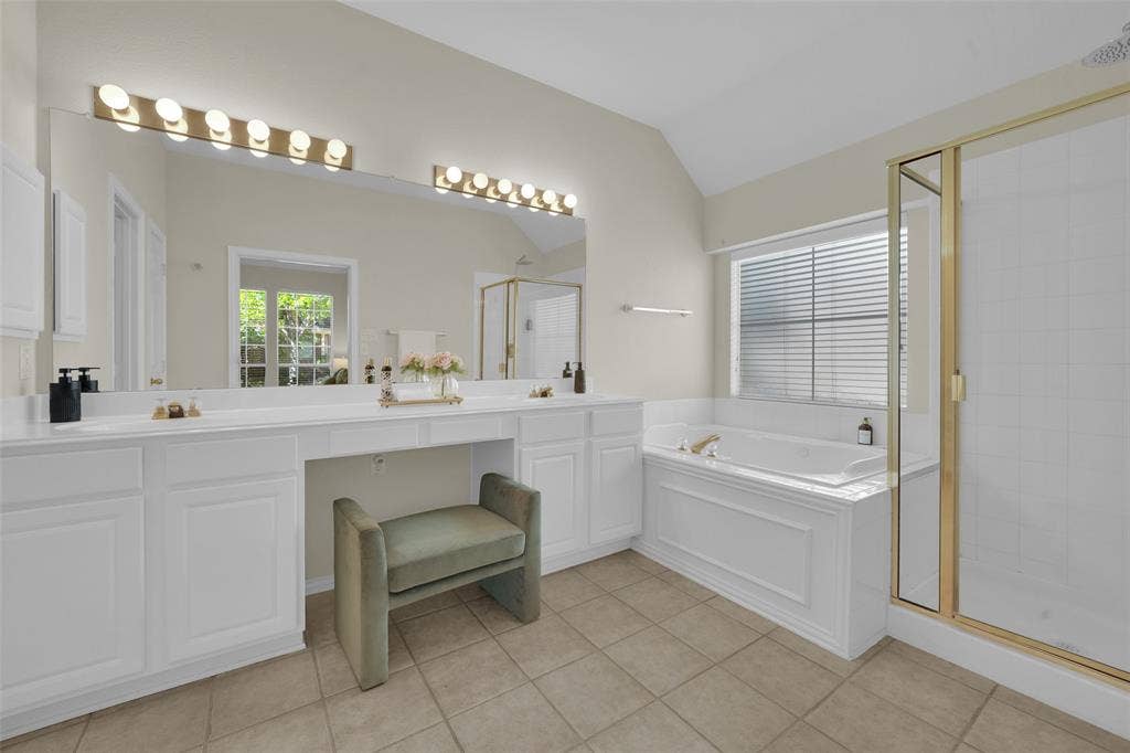 Double vanity with white cabinetry, built-in makeup counter, large vanity mirror with strip lighting, built-in soaking tub, and a glass-enclosed shower with gold-tone framing