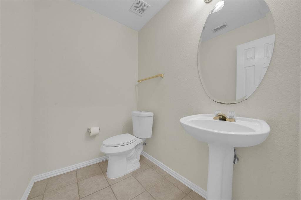 Pedestal sink with gold-tone hardware, oval wall mirror, toilet with white porcelain finish, gold-tone towel bar, and tiled flooring