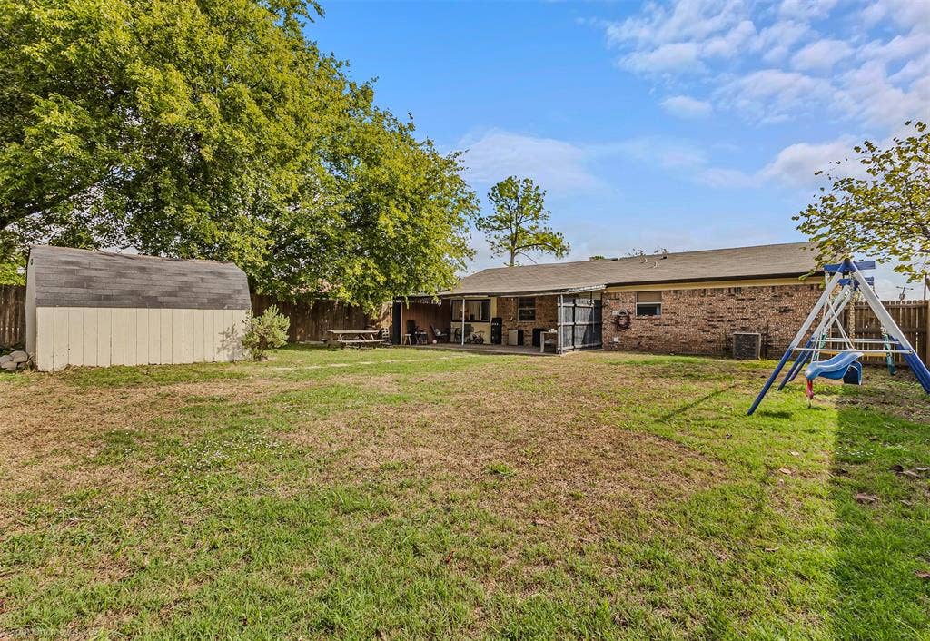 Expansive backyard featuring a brick exterior home, a covered patio, a detached storage shed, and mature perimeter trees