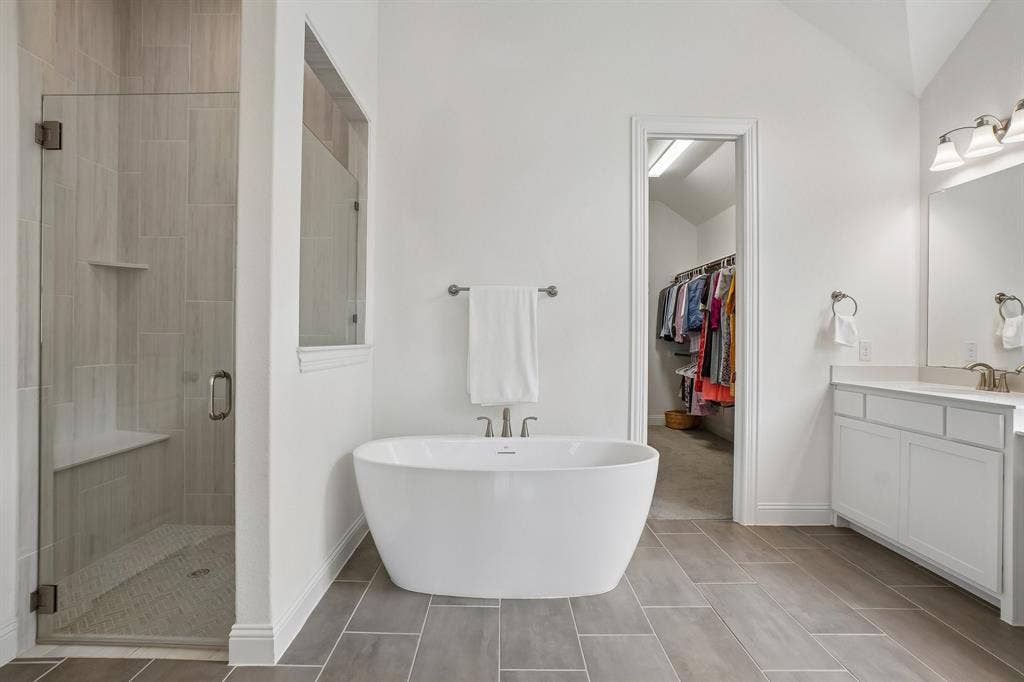 Glass-enclosed shower with a built-in bench, freestanding soaking tub, dual-sink vanity with white cabinetry, and large format floor tile