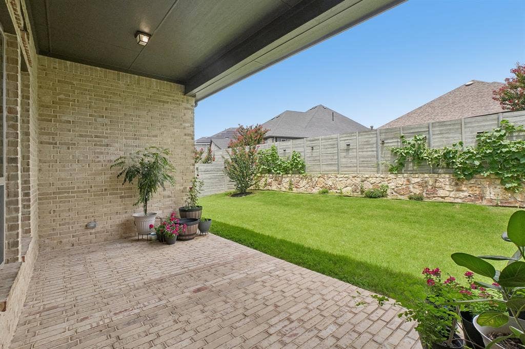 Covered outdoor patio with brick pavers, light-toned brick exterior, expansive lawn, stone retaining wall, and horizontal slat fencing
