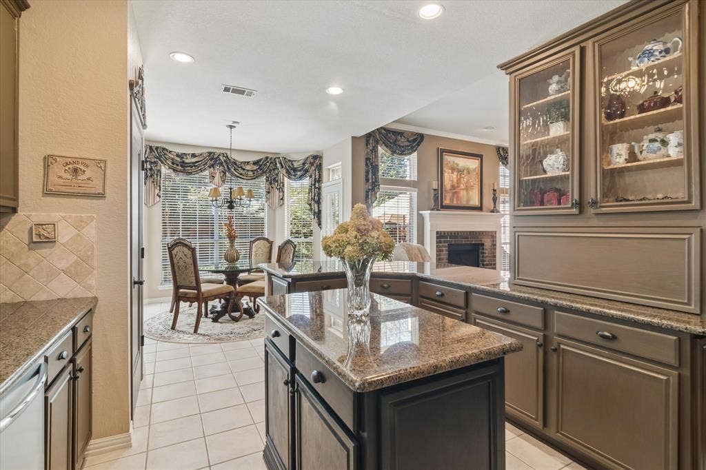 10521 Chablis Lane, Frisco, TX 75035 Kitchen island with a polished stone countertop, kitchen cabinetry with glass-front display cabinets, tiled flooring, an adjacent dining area with bay windows, and a brick fireplace visible in the background
