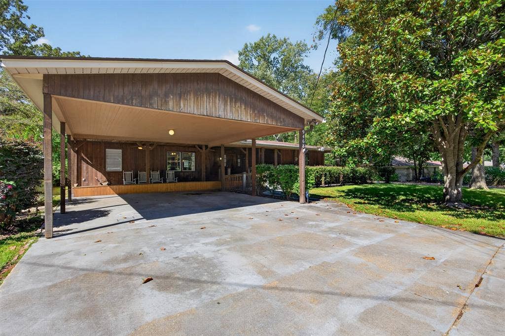 9468 Ranch Lane, Shreveport, LA 71106 Expansive concrete driveway with an attached carport, wood-paneled exterior, and mature landscaping