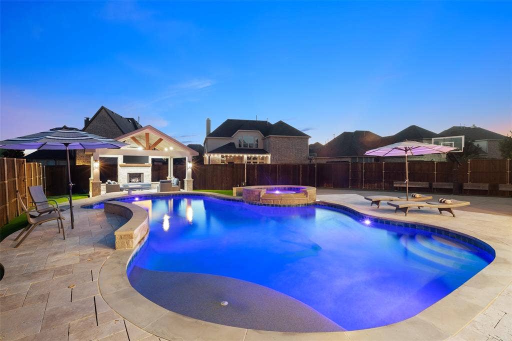 Expansive backyard featuring a custom-shaped pool with integrated spa, a built-in outdoor kitchen with fireplace, and paver stone hardscaping