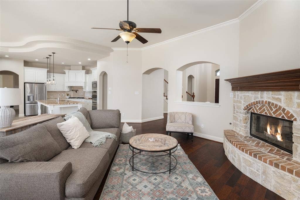 Living area featuring dark wood-finish flooring, a stone and brick fireplace with a wood mantle, and an open layout to a kitchen with white cabinetry and stainless steel appliances