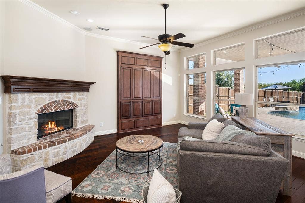 Stone and brick fireplace with a wood mantel, dark wood-finish flooring, built-in cabinetry, ceiling fan with integrated lighting, and multiple large windows overlooking a backyard pool