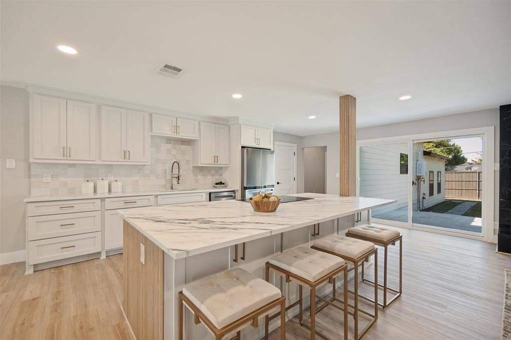 3129 Flowerdale Lane, Dallas, TX 75229 White shaker cabinetry with gold-tone hardware, a large kitchen island with a waterfall countertop, integrated stainless steel appliances, wood-finish flooring, and recessed lighting