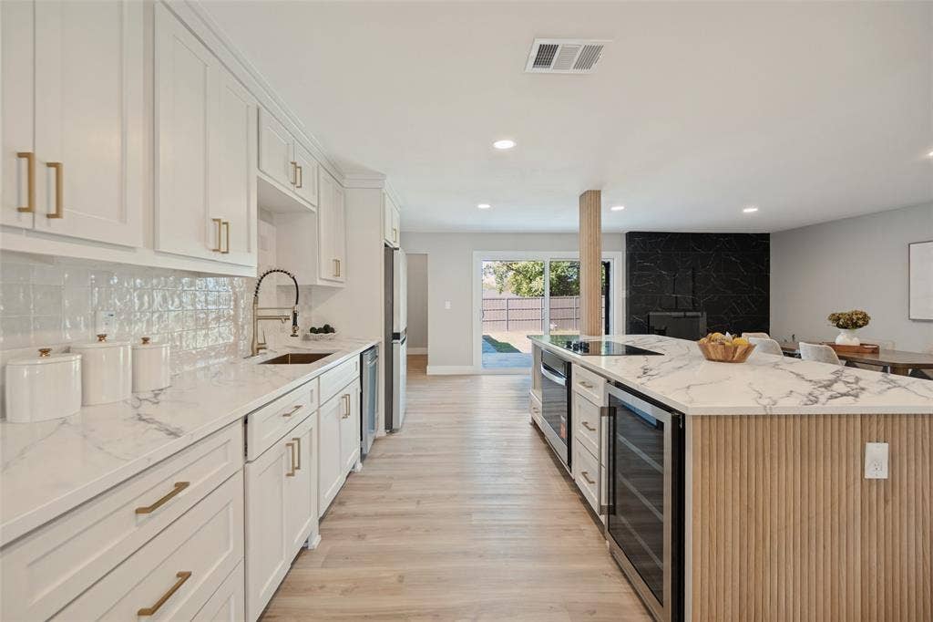 3129 Flowerdale Lane, Dallas, TX 75229 White shaker cabinetry with gold-finish hardware, white marbled countertops, light wood-finish flooring, a black marbled accent wall, and recessed lighting