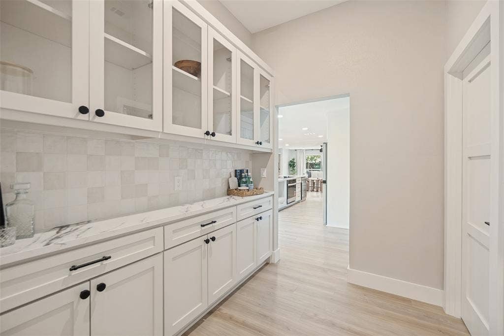 3129 Flowerdale Lane, Dallas, TX 75229 White shaker cabinetry with black hardware, glass-front upper cabinets, white tile backsplash, light wood-finish flooring, and a white countertop with gray veining