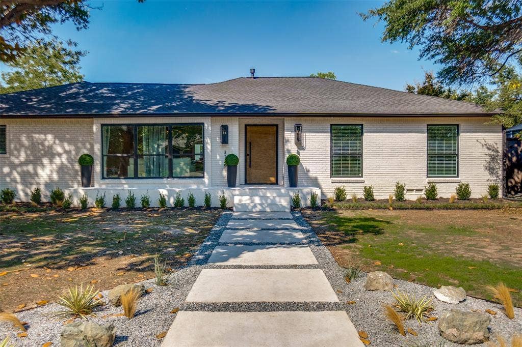 3129 Flowerdale Lane, Dallas, TX 75229 White brick exterior, black-framed windows, wood-finish entry door, concrete paver walkway, and a landscaped front yard
