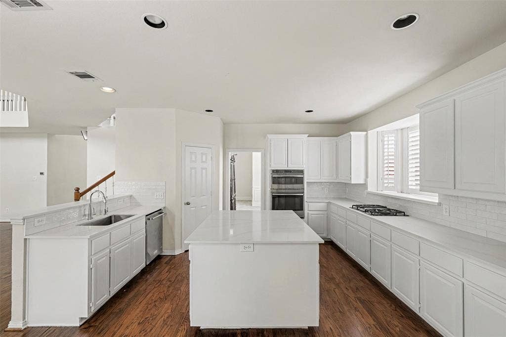7813 San Isabel Drive, Plano, TX 75025 Chef's kitchen featuring white cabinetry, a large center island with white quartz countertop, stainless steel appliances including dual wall ovens and gas cooktop, white subway tile backsplash, and wood-finish flooring throughout