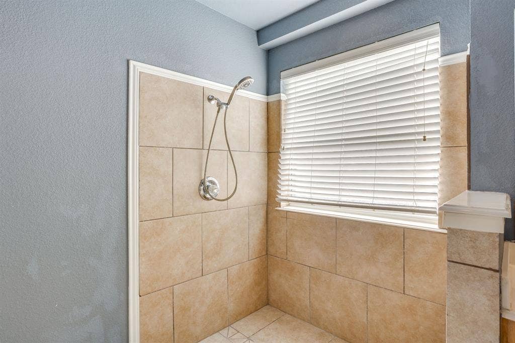 Tiled shower enclosure featuring a handheld showerhead with chrome fixtures, a window with horizontal blinds, and a tiled half-wall