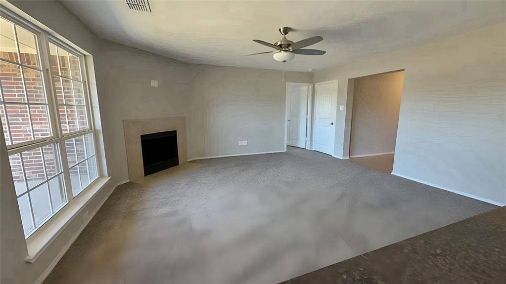 Spacious room with carpeted flooring, a corner fireplace with a light-toned surround, an overhead ceiling fan, and large multi-pane windows