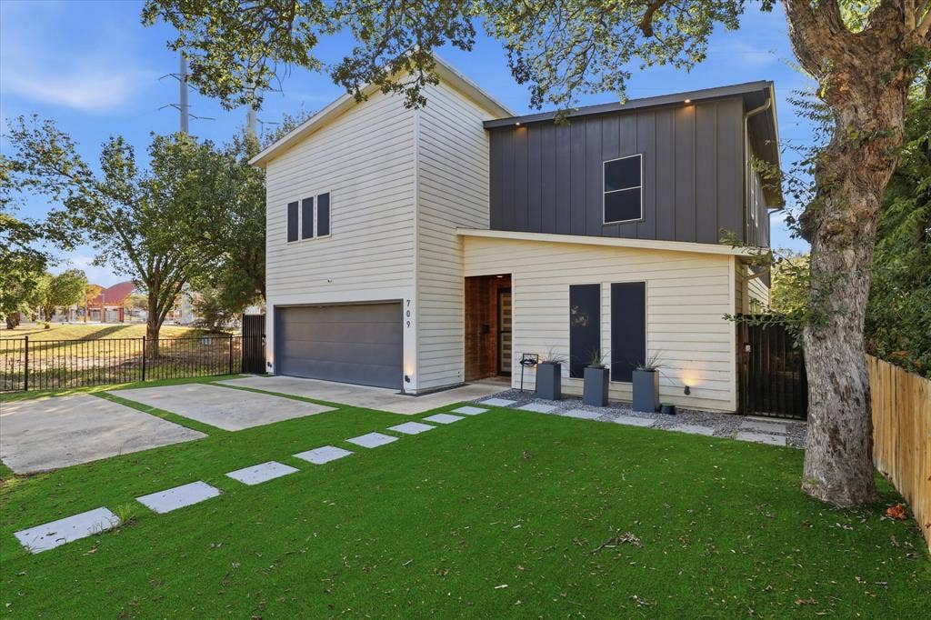 709 Woodbine Avenue, Dallas, TX 75203 Modern exterior with light siding and vertical dark metal paneling, integrated garage, concrete driveway with green accents, and a fenced perimeter