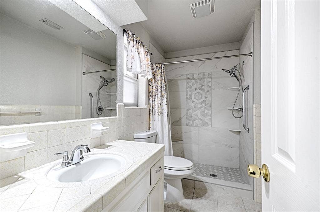 2909 Lee Park Drive, Irving, TX 75062 Bathroom featuring a vanity with a tile-surround countertop, integrated porcelain sink, full-width wall mirror, and a walk-in shower with decorative mosaic tile inlay and integrated shelving