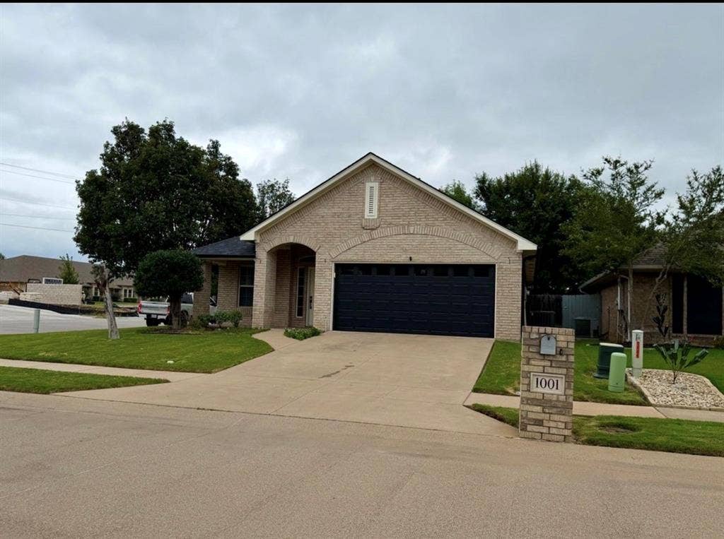 1001 Roberts Bend, Granbury, TX 76048 Brick facade home featuring a prominent arched entry, dark garage door, stone mailbox pillar, and mature landscaping