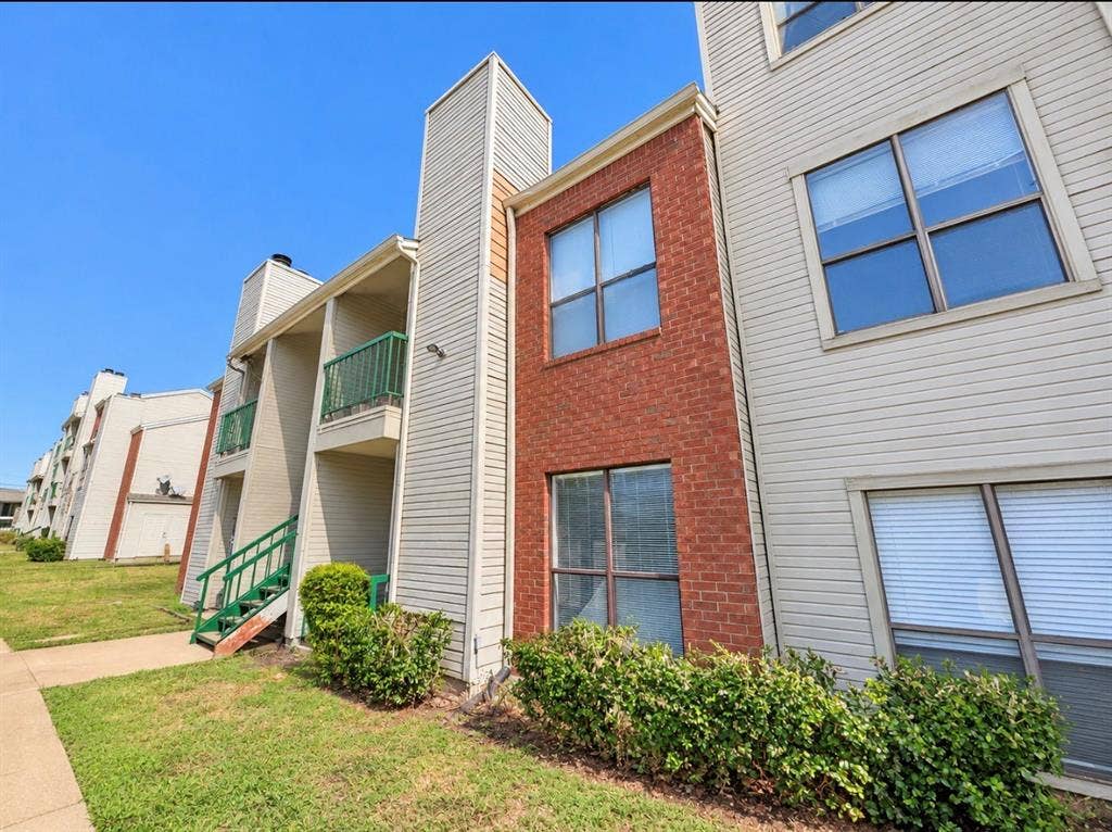 7126 Holly Hill Drive, #108, Dallas, TX 75231 Multilevel building exterior featuring light-colored siding, brick accents, multiple windows with blinds, and balconies with green railings
