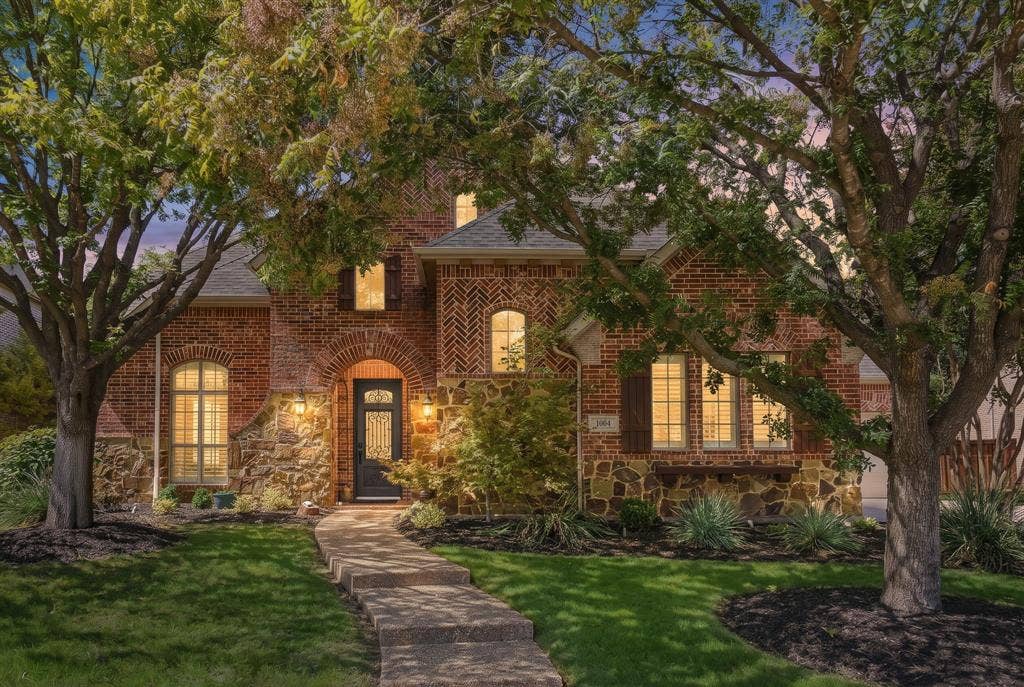 Front Exterior at Twilight | 1004 Bristlewood Drive, McKinney, Texas. Mature trees frame the illuminated facade of this five-bedroom home in Woodhaven II at Stonebridge Ranch.