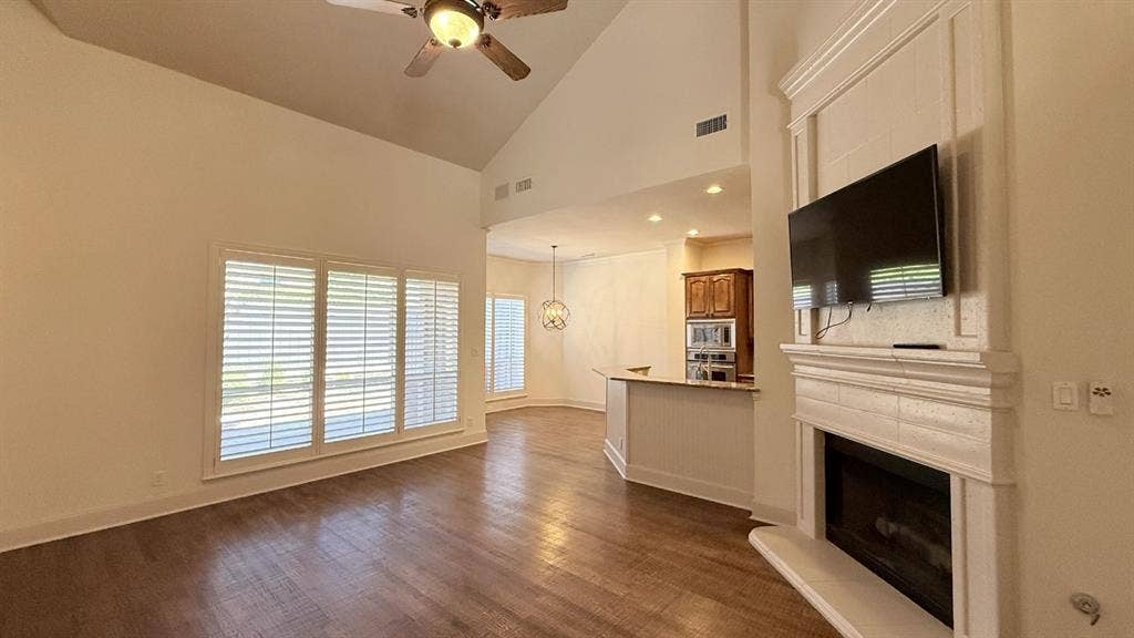 5700 Travis Drive, Frisco, TX 75034 Vaulted ceiling with a ceiling fan, wood-finish flooring, fireplace with a decorative mantel, multiple windows with plantation shutters, and a built-in kitchen island