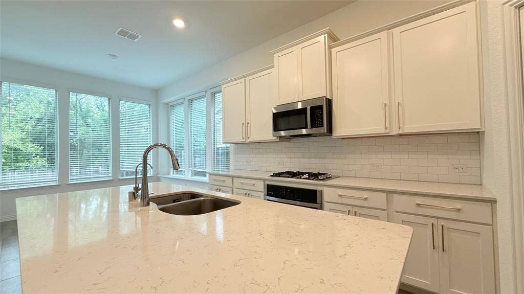 Kitchen featuring an island with an undermount sink, stainless steel appliances, white cabinetry with brushed nickel hardware, a subway tile backsplash, and large windows
