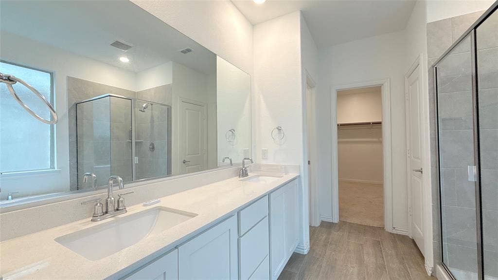 Dual undermount sinks with brushed nickel fixtures, expansive vanity with white shaker cabinetry and light-toned countertop, and a glass-enclosed shower with tile surround