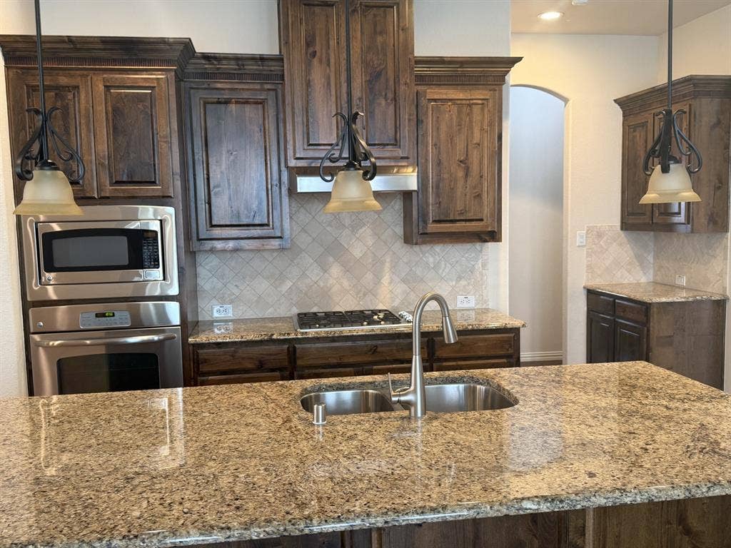 13721 Vera Cruz Road, Frisco, TX 75035 Chef's kitchen featuring custom wood cabinetry with bronze hardware, granite countertops with waterfall edge, dual stainless steel undermount sinks with gooseneck faucet, gas cooktop, and stainless steel appliances including built-in microwave and wall oven