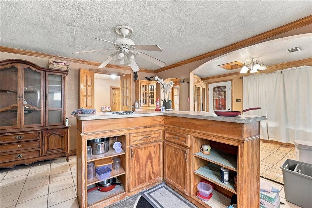 L-shaped wooden kitchen island with stone countertop, tile flooring, built-in wooden cabinetry, ceiling fan, and multi-light chandelier