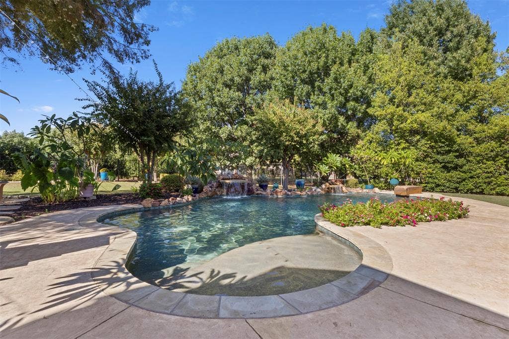 15600 Big Horn Trail, Frisco, TX 75035 Curved concrete pool deck, custom-shaped swimming pool with integrated waterfall feature, surrounding landscaping, and mature trees