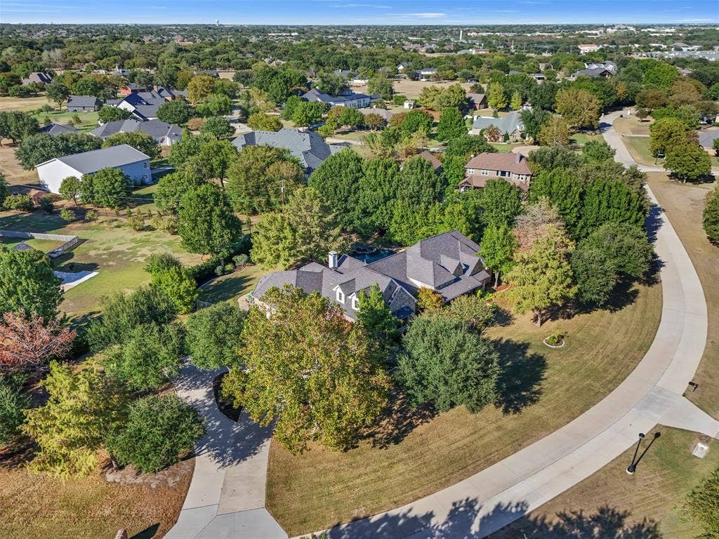 15600 Big Horn Trail, Frisco, TX 75035 Aerial perspective showcasing a property surrounded by mature trees, a curved concrete driveway, and a shingled roofline