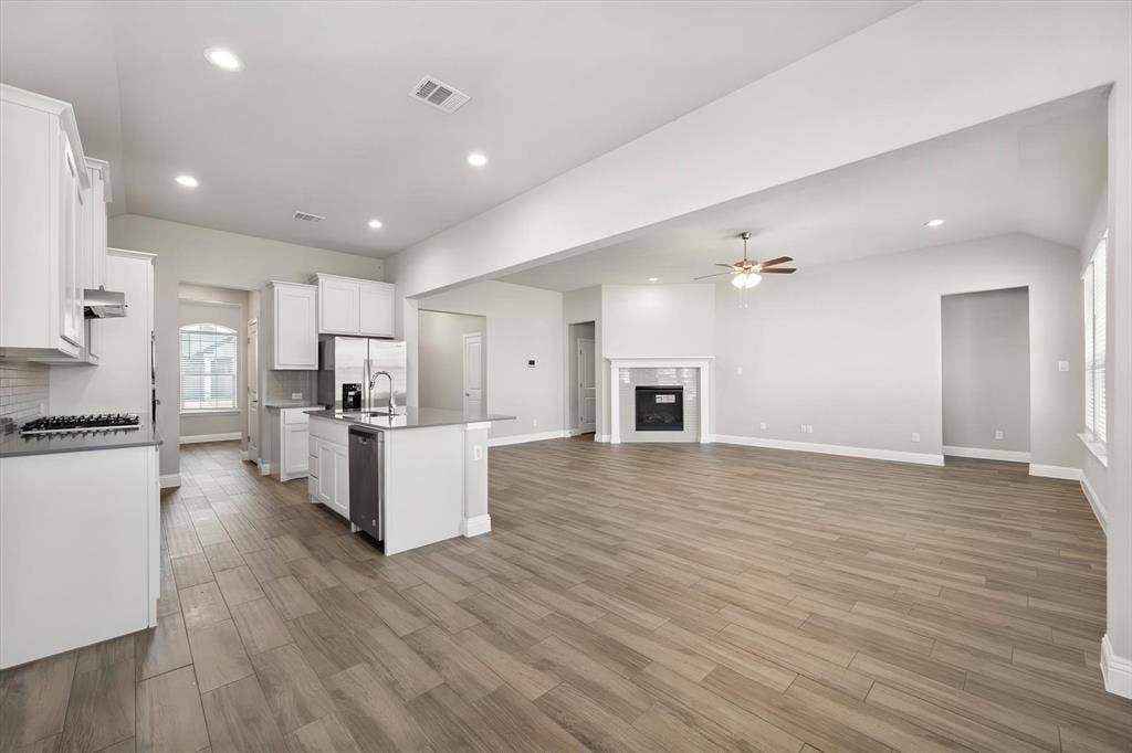 3636 Wintergreen Street, Grand Prairie, TX 76065 Open-concept living area featuring wood-finish flooring, a kitchen island with sink, white shaker cabinetry, stainless steel appliances, and a built-in fireplace with mantel