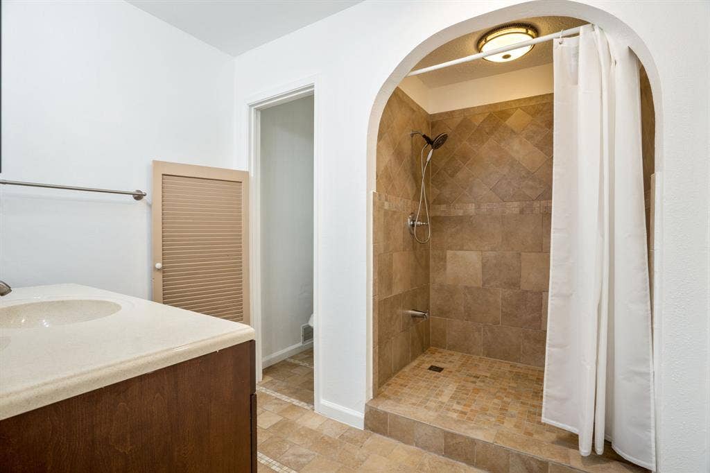 Bathroom featuring a walk-in shower with stone-finish tile surround and flooring, an arched alcove, a vanity with a solid-surface countertop, and a built-in louvered-door cabinet