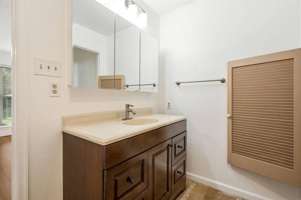 Bathroom vanity featuring stained wood cabinetry, integrated countertop with oval sink, nickel-finish faucet, tri-fold medicine cabinet, and louvered wall-mounted storage door